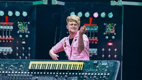 Sam Battle performing in a pink boiler suit. he is gesturing and stood next to a large keyboard with many buttons and dials on it. Large electronic equipment is visible in the background as stage decoration.