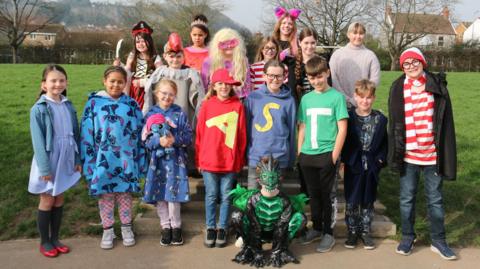 A large group of children dressed in various outfits for World Book Day stand outside on the playing fields. Some of them are dressed as "Where's Wally" and others are dressed as pirates and animals