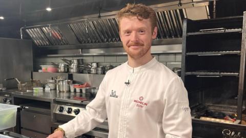 Ewan Waller is standing in the a restaurant kitchen, with pots and pans behind him. He smiles into the camera and wears a chefs apron that says 'Michelin 2026.'