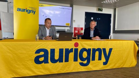 Two men sitting behind a table with a yellow banner with the word Aurigny on it. 