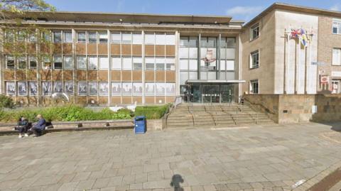 A large brick building with dozens of windows on the front. Flagpoles, benches and bushes are outside the building.