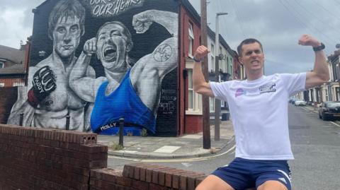 Joe Wild during his marathon in Liverpool, posing with his biceps flexed in front of a mural of martial arts fighters Paddy Pimblett and Molly McCann.