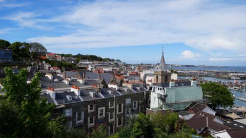 A view over St Peter Port in Guernsey. A row of houses and a church sit among trees, with a harbour and sea behind.