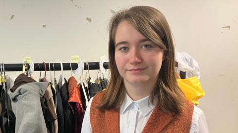 A woman in a white shirt and an orange waistcoat stands in front of a clothing rail with lots of clothing hanging on it.