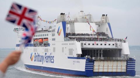 A white Brittany Ferries with blue writing and highlights, against a grey sky with a blurry Union Flag being waved in the foreground.