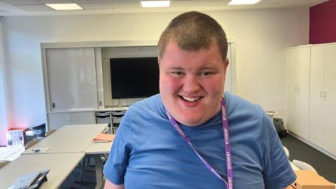 Photograph of Sam from Heald Green in Stockport. The image is taken inside one of the classrooms at Royal College Manchester. Sam is smiling and wears a blue t-shirt with a purple "student" lanyard.