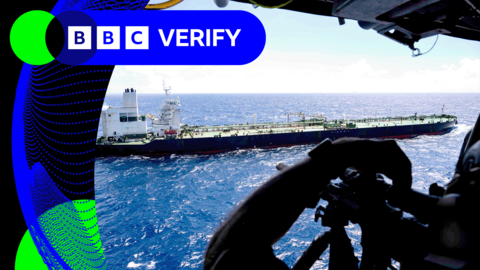 A picture of a tanker sailing in the ocean taken from a military helicopter, you can see a soldier aiming a rifle in the foreground 