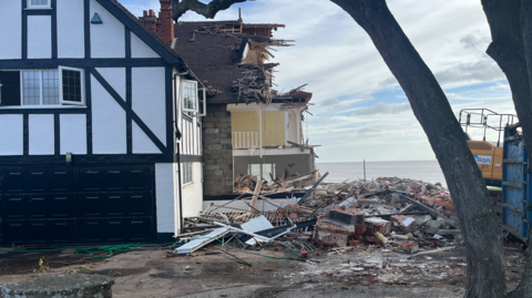 A partially demolished home on the coastline next to a pile of debris. In the background is the sea, blue sky and white clouds.