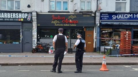 Two police officers in uniform stand on a road in front of police tape which surrounds shops and restaurants. Their backs are to the camera. Signage for the businesses pictured indicate they are a barbershop, an Indian restaurant and a deli.
