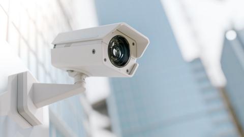 A CCTV camera on a wall with blurred buildings in the background