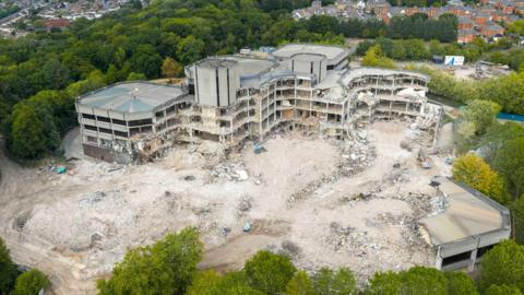 A destroyed area of land and buildings next to three remaining buildings. It is surrounded by trees and in the distance there is a housing estate.