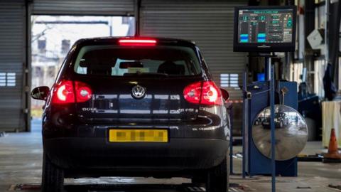 A black car is parked beside a computer which is doing the MOT test.