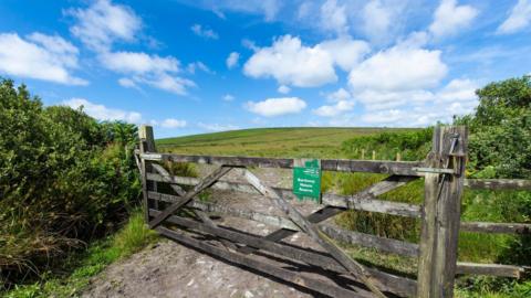 A large gate is seen closing off a huge verdant field with shrubbery to each side. A sign reads Bartinney Nature Reserve 