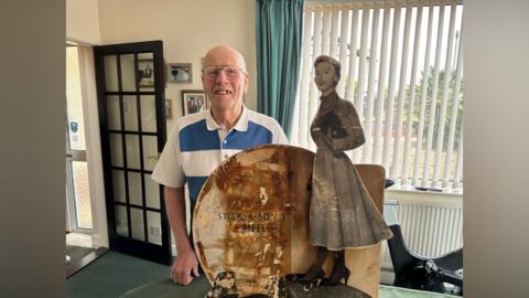 A smiling Reginald Wright looks directly at the camera as he stands in his living room behind the sign. The sign has a woman in high heels and a midi-length dress standing and carrying a clutch handbag. 