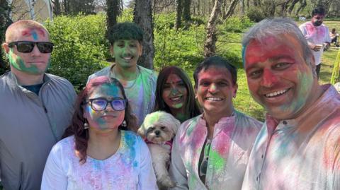 A group of six people - four men and two women - and a dog. They are looking at the camera and smiling. They are all covered in brightly coloured powders of various colours. 