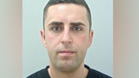 Joshua Adcroft's mugshot. He has short dark hair and is wearing a black t-shirt against a plain grey background