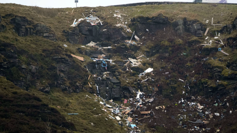 Waste scattered down a mountain with a lay-by above. It's not possible to pick out individual items but there are sheets of wood and small items covering the rocky ground.