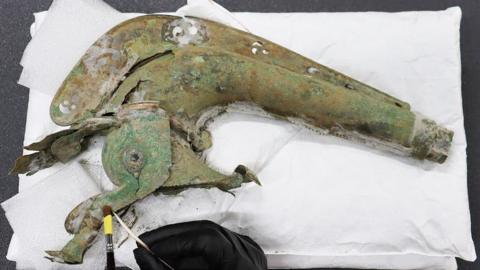 The head of an Iron Age carnyx resting on its side on a white pillow. The instrument is in the shape of an animal with a long neck, with a crest along its neck, pierced with a pattern, with a long nose and its mouth gaping open. A black gloved hand can just be seen at the bottom holding a very fine brush, while a much broader brush brushes the snout of the animal.