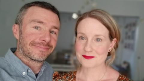 A close-up photo of Danny Riordan and his wife Maria Moynihan looking to camera and smiling.  Danny has short hair and a beard and is wearing an open-neck grey shirt. Maria has blonde hair swept back from her face.  She is wearing a floral v-neck top or dress with a gold necklace, earrings and red lipstick.