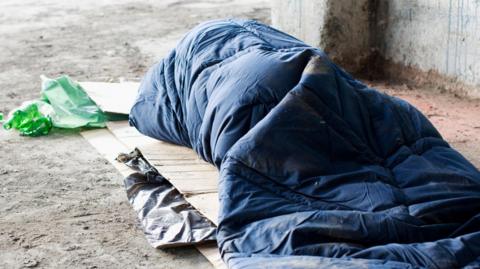 A homeless person is inside a sleeping bag. They are outside and on top of cardboard.