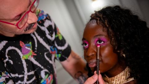 A young black woman has pink eye shadow applied to her face with a brush. The man applying it has a moustache and red glasses