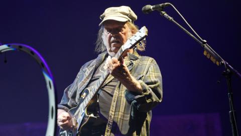 A man standing onstage playing a guitar. He is wearing a grey hat and a denim top. 