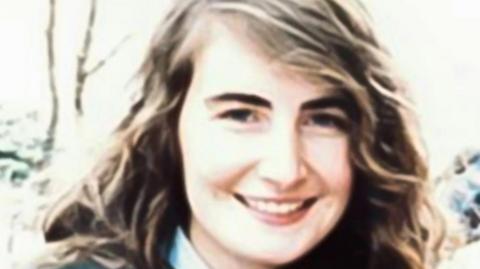 Annie McCarrick has long wavy brown hair with hints of blonde.She is wearing what appears to be a blue shirt and navy jacket. 
She is smiling and can be seen standing outside. 
A number of trees and bushes can be seen behind her. 
