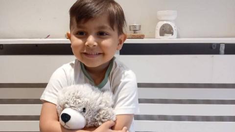 Zak in a white t-shirt is cuddling his white teddy bear with a big grin on his face