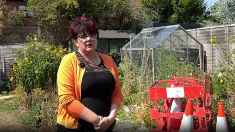 Melissa Stroud standing in front of a manhole with traffic cones and barriers around it. There are plants around a greenhouse behind it.