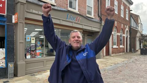 A man with short grey hair smiling while he puts both his hands in the air with his fists clenched. He is wearing a blue jacket and a blue top. Behind him is a row of buildings including one that has "LincIT" on the its sign.