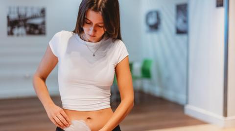 A young woman in a cropped white t-shirt is standing in a room holding an ostomy bag. She is looking down towards it and placing it in her trousers. 