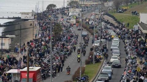 Hundreds, if not thousands, of motorcycles and people lining a seafront road as more motorcycles drive along it.