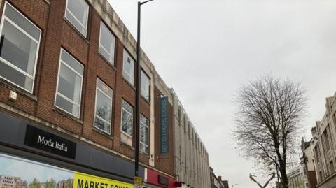 A red brick building with a sign underneath saying "moda italia". Another sign in the distance reads "British Home Stores". A grey concrete building is next door.