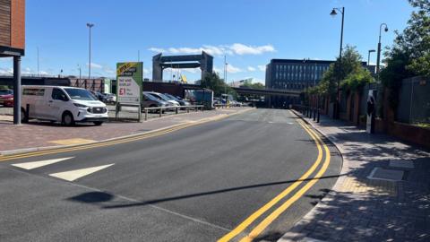 A street with double yellow lines runs past a car park which has a large green and red sign. Several cars are parked on the site, the Hull tidal barrier and modern buildings are visible in the background.