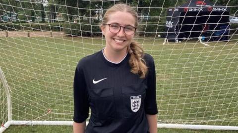 A teenage girl with blonde hair in two plaits, wearing glasses and a black referee uniform, stood in front of a goal on a football pitch