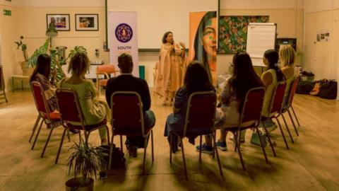 South Asian Voices Bristol CIC hosting a group discussion in a function room in Bristol. One member is speaking using a microphone. Seven other people are listening while seated in a semi circle. The woman speaking is wearing a sari. 