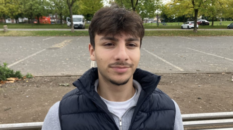 A young man with dark hair, a moustache and goatee style beard. He has a blue bodywarmer over a grey top and white tshirt. There is a car park behind him