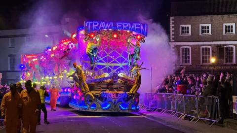 An illuminated carnival float drives down the road at night, as groups of people watch from the street