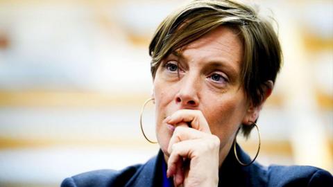 Jess Phillips looks on during a meeting as she wears a dark blue jacket. She has short, light brown hair and gold hooped earrings.
