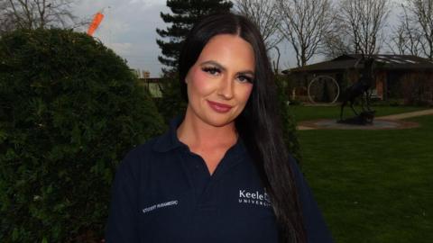A woman with long black hair and wearing a blue shirt which says "student paramedic" and "Keele University", smiles while standing outside.
