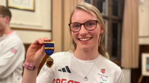 Rebecca Redfern pictured with large black frame glasses. Her hair is in a blonde ponytail and she is smiling towards the camera. She is wearing a white Great Britain t-shirt, silver necklace and pink watch while holding a small golden medal with a blue ribbon on it.