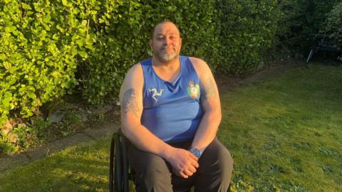 Mike Ryan, a man with tattoos on his arms, he wears a blue vest with the three legs of man and smiles as he sits in the garden in his wheelchair.