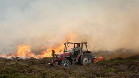 A farmer drives his tractor across heathland while a wildfire burns in the background