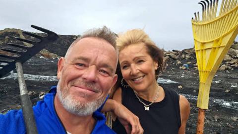 Dean Ward and Allie Evans. He has grey hair and a beard and is wearing a blue top. She has a black top, silver earrings and a silver necklace with butterfly pendant. They are holding rakes and stood on a blackened mountain