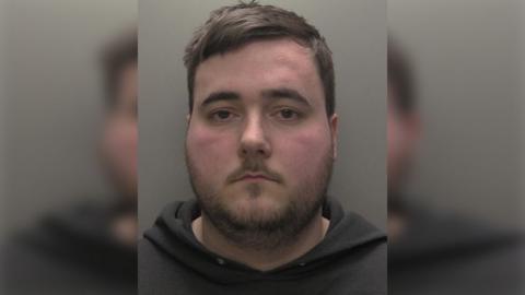 Custody photograph of Connor Laud he has short brown hair and beard and is wearing a dark hooded top