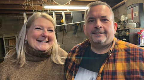 A woman with shoulder-length blonde hair in a brown polo neck and a man with short grey hair in a checked shirt smile at the camera