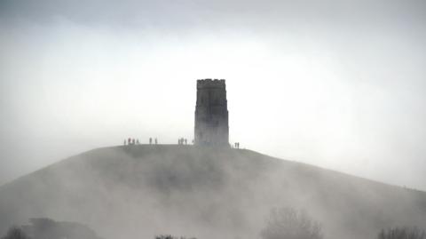 Glastonbury Tor in the fog. Silhouettes of walkers can be seen around St Michael's Tower.

