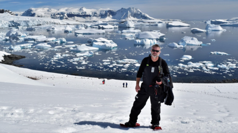 Man standing on snow in black outfit with snow-covered water in background behind him