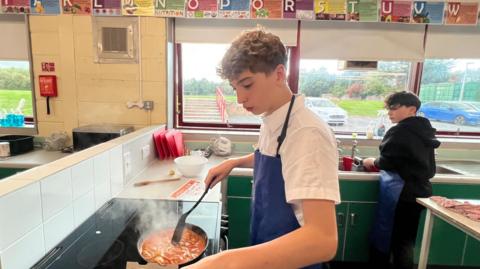A school boy cooking chicken tikka masala at a stove. He is stirring a pot and is wearing a blue apron. He is looking down at the food, which is steaming hot. Another boy is in the background washing dishes.