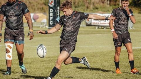 A young rugby player in a black strip prepares to kick the ball in a game